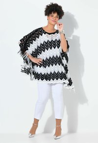 Svartvit zigzag-mönstrad poncho med fransiga kanter, bärs över vita sittande byxor och silverfärgade klackar. Texturerat stickat material.