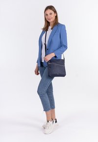 Borsa a tracolla in pelle blu navy con superficie strutturata, tasca frontale con zip e tracolla regolabile. Abbinata a un blazer blu e denim chiaro.