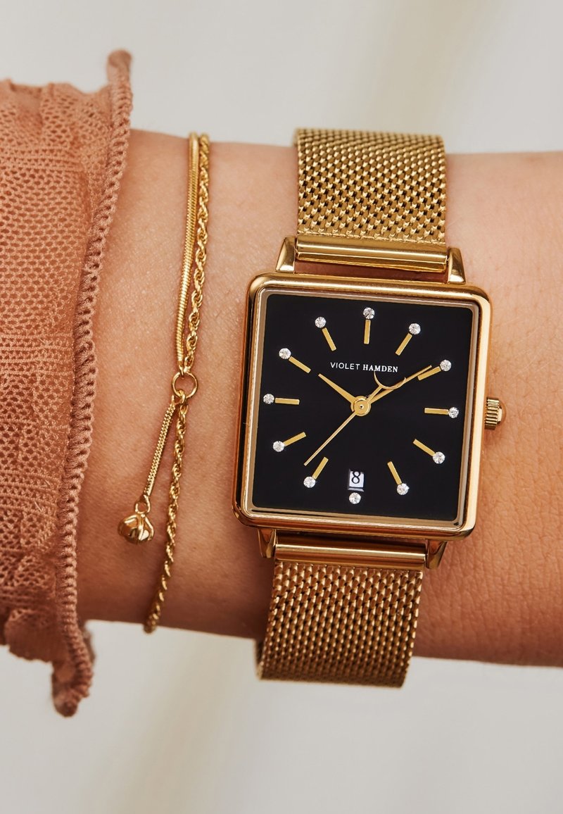 Montre avec bracelet en maille dorée, dotée d'un cadran carré noir, de repères horaires dorés et d'accents argentés. Comprend un bracelet délicat avec un petit pendant.