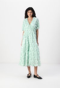 Long, mint green dress with a V-neckline. Features layered fringe detailing, short sleeves, and a cinched waist. Black flat shoes.