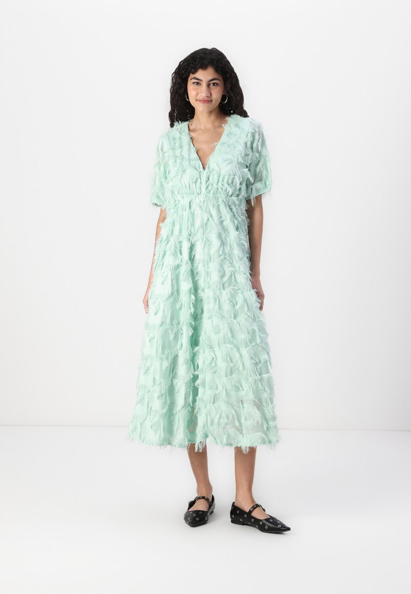 Long, mint green dress with a V-neckline. Features layered fringe detailing, short sleeves, and a cinched waist. Black flat shoes.