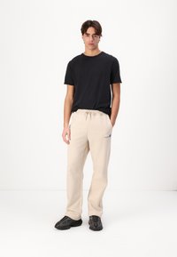 Schwarzes T-Shirt mit kurzen Ärmeln kombiniert mit beigen Jogginghosen mit Kordelzug, präsentiert von einem männlichen Model mit schwarzen Sneakers.