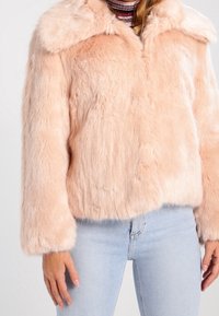 Kvinna i ljusrosa fuskpälsjacka över randig polo och ljusblå jeans, står med armarna avslappnat vid sidorna.