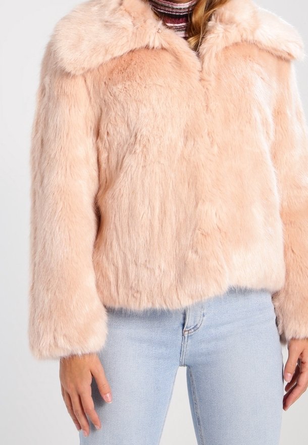 Kvinna i ljusrosa fuskpälsjacka över randig polo och ljusblå jeans, står med armarna avslappnat vid sidorna.