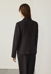 Černé sako na míru s výrazným střihem, zapínáním na jeden knoflík a minimalistickým stylem. Materiál vypadá hladce s jemným leskem.