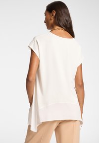 Cremefarvet top med korte cap sleeves, en afslappet pasform og et lagdelt kant. Har en glat tekstur og en rund halsudskæring.