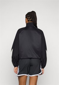 Svart satengjakke med høy krage og ballongslem, med hvite detaljer på skuldrene. Kombinert med svarte shorts.