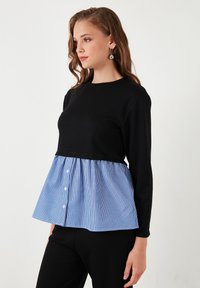 Top nero a maniche lunghe con un peplum a righe blu e bianche. Presenta un colletto rotondo e dettagli con bottoni sul peplum.