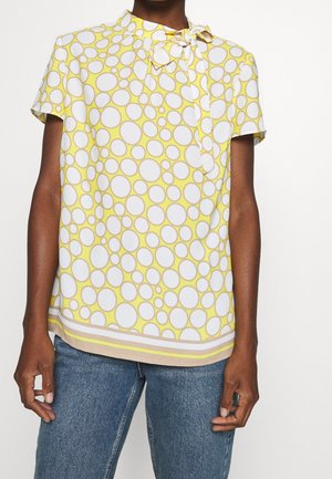 Persona que lleva una blusa de manga corta amarilla con un patrón de círculos grandes en blanco y beige y un lazo en el escote, combinada con vaqueros azules.