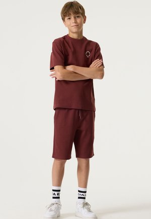 Bordeauxrode crewneck T-shirt met logo, gecombineerd met bijpassende shorts. Witte sneakers en zwart-witte sokken met patroon maken de outfit compleet.