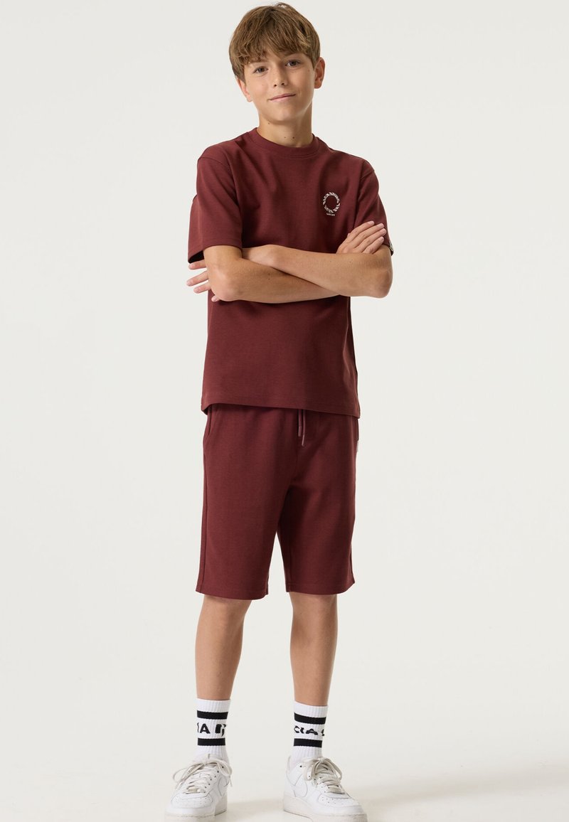 T-shirt col rond bordeaux avec logo, assorti à un short assorti. Des baskets blanches et des chaussettes noires et blanches à motifs complètent la tenue.