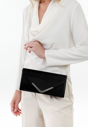Femme portant une tenue blanche tenant une pochette noire style enveloppe avec un fermoir argenté en forme de V et une bandoulière en chaîne portée en travers du corps.