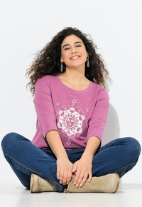 Lila Langarm-T-Shirt mit einem weißen kreisförmigen Grafikdesign. Blaue Jeans und beige Knöchelstiefeletten. Model sitzt auf dem Boden und lächelt.