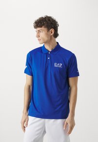 Blå poloshirt laget av glatt stoff. Har krage, korte ermer, to knapper, og en brodert "EA7"-logo på brystet.