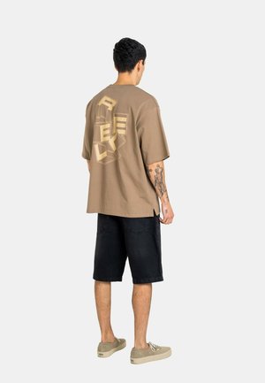 Mann mit Arm-Tattoo trägt übergroßes braunes T-Shirt mit "REVO" Blockbuchstaben auf dem Rücken, schwarze Shorts und beige Sneaker, steht mit dem Rücken zum Betrachter.