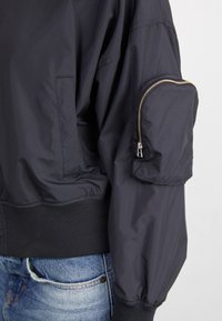 Zwarte, lichte bomberjack met een ritszak op de bovenarm, ribgebreide zoom en manchetten, een gladde textuur en een losse pasvorm.