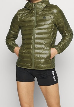 Persona che indossa una giacca imbottita Adidas verde oliva con cappuccio e pantaloni corti sportivi neri Terrex, con le mani nelle tasche della giacca, in piedi.