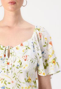 Blusa floreale bianca con maniche puff corte, scollatura quadrata e dettaglio a fiocco davanti, caratterizzata da vivaci motivi floreali.