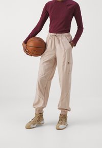 Beige baggy broek met een elastische tailleband en boorden, voorzien van zijzakken en gestikte details. Gepaarde met een kastanjebruine lange mouw top.