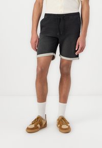 Shorts en denim noir avec une coupe décontractée, dot�és d'une taille élastique à cordon et de bords roulés. Portés avec des baskets beiges et marron et des chaussettes blanches.