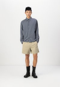 Camisa gris de botones, pantalones cortos beige y botas negras hasta los tobillos. La camisa tiene un ajuste relajado y los pantalones cortos son de longitud hasta la rodilla.