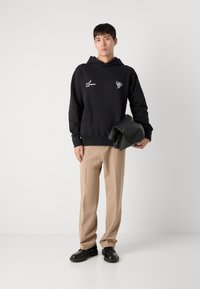 Sweatshirt à capuche noir avec poche kangourou à l'avant, arborant des impressions graphiques blanches. Assorti à un pantalon beige clair et des chaussures noires.