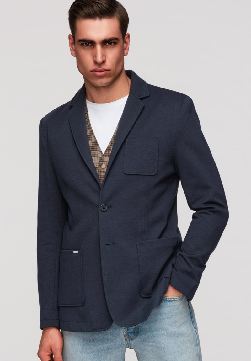 Junger Mann mit marineblauem Blazer über brauner Weste und weißem Hemd, Hände in den Taschen, vor schlichtem hellgrauem Hintergrund.