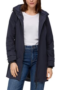 Marineblauwe waterdichte jas met capuchon, voorrits en lange mouwen. Voorzien van manchetdetails en een relaxte pasvorm, gecombineerd met een wit shirt en jeans.