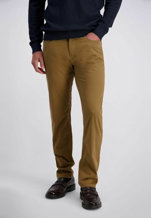 Mann trägt senfgelbe Hose, schwarzen Pullover und dunkelbraune Leder-Loafer, steht mit einer Hand in der Tasche vor einfarbigem Hintergrund.