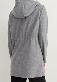 Manteau gris à capuche avec taille cintrée, manches longues, et longueur mi-cuisse porté sur un pantalon noir, vu de dos.
