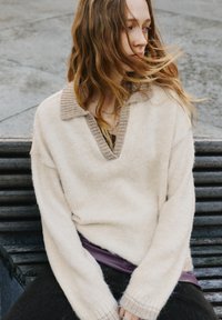 Beiger Pullover mit V-Ausschnitt und geripptem Kragen, aus weichem Material gefertigt. Verfügt über überschnittene Schultern und lange Ärmel mit gerippten Bündchen.