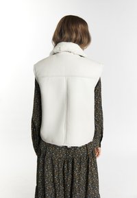 Witte leren mouwloze vest met een bontkraag, gladde textuur en strakke lijnen, gedragen over een donker bloemenpatroon jurk.
