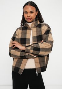 Braun- und schwarzkariertes Zip-Jacke mit hohem Kragen, Reißverschluss vorne und langen Ärmeln, kombiniert mit einem beigen Pullover darunter.