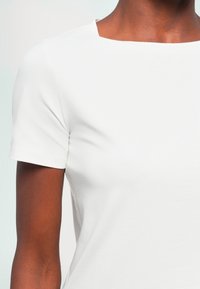 Gros plan d'une personne portant une chemise blanche à manches courtes avec un large col arrondi sur un fond clair.