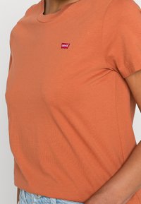Camiseta de algodón de color óxido con mangas cortas. Presenta un pequeño logo rojo de Levi's en el pecho. Diseño ajustado con cuello redondo.