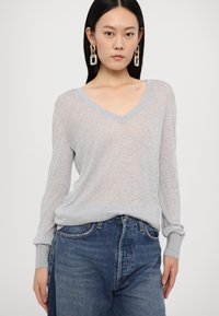 Pull à col en V gris clair à manches longues avec une texture douce, présentant une coupe décontractée et des manches retroussées, associé à un jean bleu taille haute.