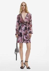 Blumenmusterkleid in Rosa- und Lila-Tönen, mit tiefem V-Ausschnitt, langen Ärmeln und gebundenem Taillenbereich. Stylisch kombiniert mit schwarzen Riemchen-Absätzen und einer grauen Tasche.