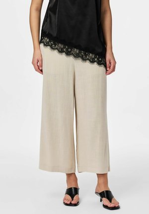 Kvinne iført beige vide, korte bukser, svart topp med blonder, og svarte støvler med åpen tå og hæl, stående foran en hvit bakgrunn.