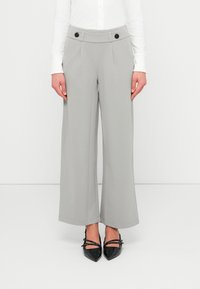 Γυναίκα που φοράει ανοιχτόγκρι παντελόνι με φαρδιά μπατζάκια και μαύρα κουμπιά στη μέση, καθώς και μαύρες μυτερές γόβες Mary Jane, στέκεται μπροστά σε λευκό φόντο.