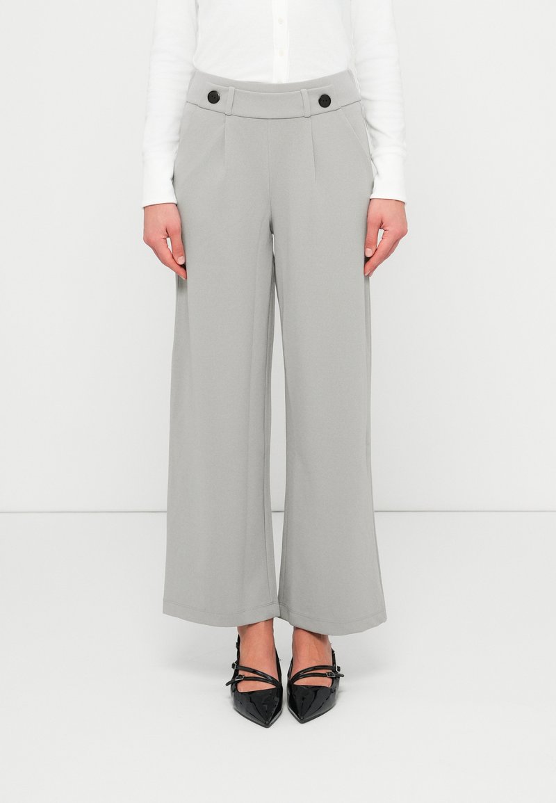 Γυναίκα που φοράει ανοιχτόγκρι παντελόνι με φαρδιά μπατζάκια και μαύρα κουμπιά στη μέση, καθώς και μαύρες μυτερές γόβες Mary Jane, στέκεται μπροστά σε λευκό φόντο.