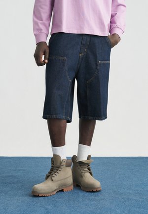 Μαύρο άτομο που φοράει σκούρο τζιν σορτς, ταμπά μπότες από σουέντ με λευκές κάλτσες και ένα ανοιχτό μοβ μακρυμάνικο πουκάμισο, στέκεται πάνω σε μπλε χαλί.