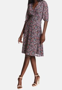 Květinové zavinovací šaty s krátkými balónovými rukávy, tmavé pozadí, růžové a bílé květy, po kolena s upnutým pasem, doplněné tělovými vysokými podpatky.