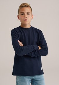 Jūrinės mėlynos spalvos ilgomis rankovėmis medvilninė marškinėlių palaidinė su atviru raštu, apvalia iškirpte, šoninėmis skeltelėmis ir maža prekės ženklo etikete kairėje apačioje.