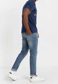 Marineblaues T-Shirt mit kurzen Ärmeln und weißem Grafikdesign, kombiniert mit hellblauen Slim-Fit-Jeans. Das Model trägt weiße Sneakers mit hochgerollten Säumen.