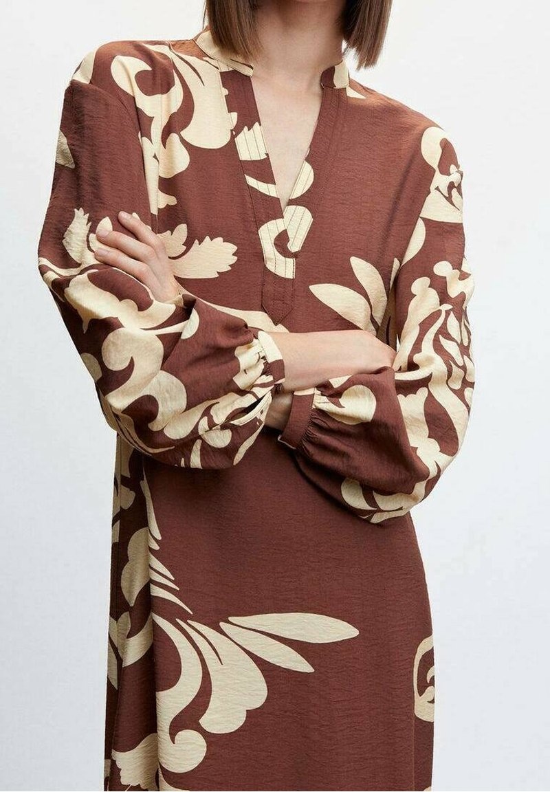 Femme les bras croisés portant une robe marron avec de grands motifs floraux crème et de longues manches bouffantes.