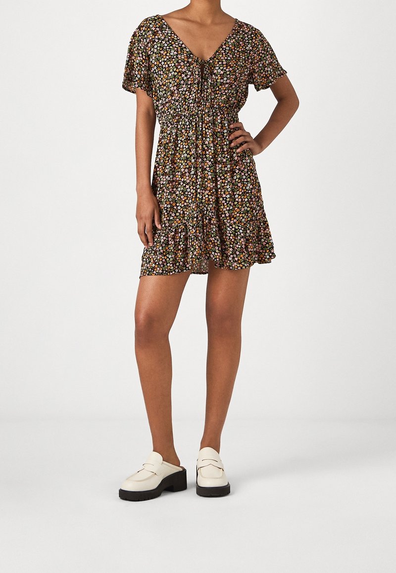 Kurzes Kleid mit floralem Muster, V-Ausschnitt und elastischem Bund, ausgestattet mit kurzen Ärmeln und einem Rüschensaum. Getragen mit weißen Loafers.