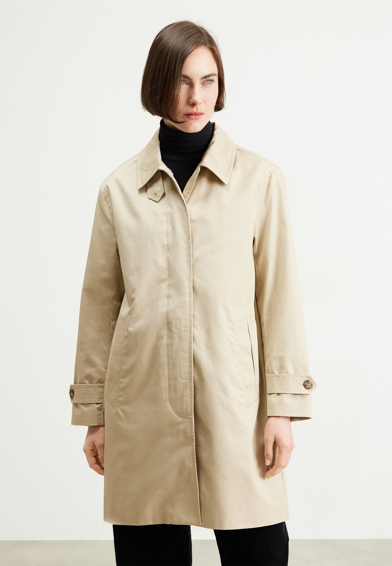 Abrigo beige con cuello clásico, puños abotonados y bolsillos laterales. Textura suave y diseño recto.