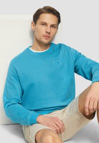 Türkiser Baumwollpullover mit Rundhalsausschnitt und gerippten Bündchen, kombiniert mit hellbeigen Shorts. Glatte Textur, minimale Markenkennzeichnung auf der Brust.