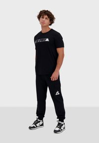 T-shirt noir à manches courtes avec un logo blanc, associé à un pantalon cargo noir avec une poche latérale et des accents blancs, complété par des baskets noires et blanches.