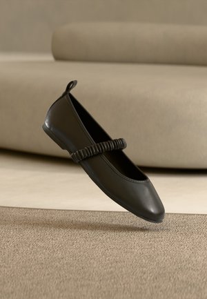 Scarpa piatta nera con cinturino elastico arricciato sulla parte superiore, mostrata sopra moquette beige texturizzata e sullo sfondo di un divano beige chiaro.